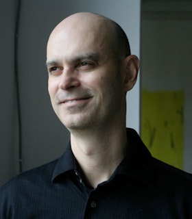 A portrait of Arturo Herrera looking to the left in front of a white wall. He wears a black collar shirt and slightly smiles.