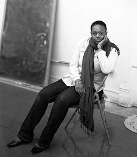 A black and white portrait of Leslie Hewitt sitting sideways on a folding chair. A large blackboard with some smudged chalk drawings rests on the wall behind her. She has short black hair and wears a white button up shirt, dark jeans, black ballet flats, and a long dark scarf. She looks towards the camera and rests her chin in one hand. 