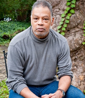 A portrait of Ishmael Houston-Jones sitting on a chair in front of trees with bright green foliage. He has short grey hair and wears a grey turtle neck, blue jeans, and a watch with a brown leather strap. His hands are clasped in his lap. 