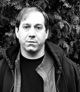 A black and white portrait of Matt Hoyt in front of a pine tree. He has short hair and wears a black unzipped hoodie, a grey cardigan, and a black shirt. 