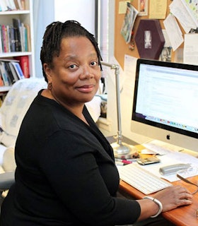 Erica Hunt wears a black top and sits at a desk computer. She looks directly at the camera and smiles slightly. In the background, there is the a full bookshelf, a bulletin board, and a white arm chair with a quilt over it.