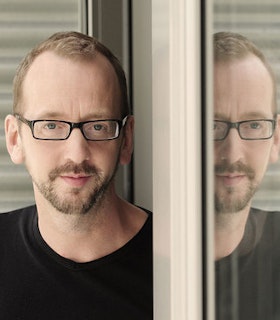 A portrait of John Jasperse standing beside a mirror and reflected in it. He has short copper hair and wears thin rectangular black glasses, a black shirt.