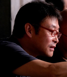A close up, side angled portrait of Jianwei Wang conferring with another person who is blurred along with the rest of the background. Wang has short black hair and wears a navy t-shirt and red glasses. 