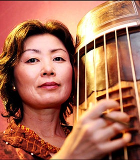 Jin Hi Kim faces the camera as she plays her komungo. She has shoulder length black hair and wears an orange shirt. The background is pink.