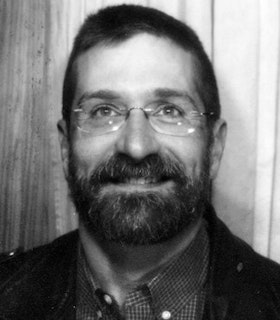 A black and white portrait of William E Jones in front of a wood wall. He has short hair and wears rimless glasses, a checkered button up shirt and a suit jacket.