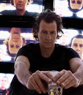 Jon Kessler sits in front of projections of his own face at different angles. He wears all black and looks intently at a piece of equipment in his hands.