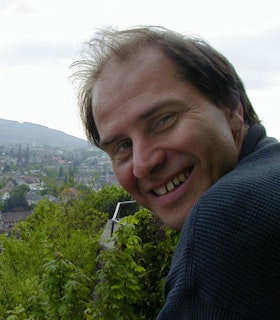 A portrait of Ron Kuivila in front of tree lined city scape. He has short brown hair, wears a blue sweater and is turning towards the camera, smiling. 
