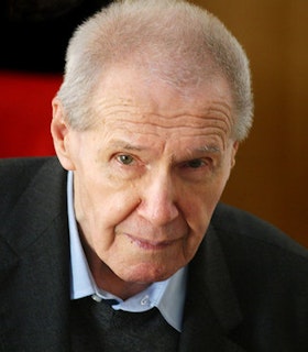 A close up portrait of Gyorgy Kurtag in front of a blurred red background. He smiles slightly and looks at the camera. He has short white hair and wears a light blue collared shirt and a dark sweater.