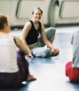 A portrait of Jennifer Lacey sitting on the floor of a dance studio in front of people. She wears a black tank top and grey sweatpants and smiles as she looks towards the camera. 