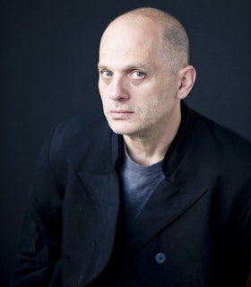 A three-quarter profile portrait of David Lang wearing a dark suit jacket and a grey t-shirt in front of a dark background. He looks directly at the camera.