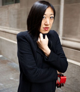 A side angle portrait of Young Jean Lee in front of a beige color building. She clutches her black jacket with her left hand while holding a red double-row tambourine on her right hand.