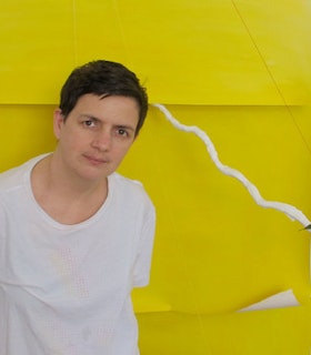 A portrait of Siobhan Liddell wearing a white t-shirt and standing in front of a bright yellow background made of paper which curls up at the edges. Liddell looks directly at the camera with a peaceful expression on her face. 