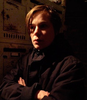 A side angle portrait of Klara Liden in a dark lit room with shadows across the left side of her face. She has short blonde hair which is swept to the side and wears a black sweater. Her arms and crossed in front of her chest. Behind her, there is a rusted white wall with many panels.  