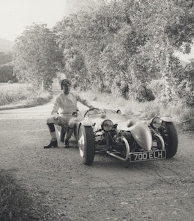 A black and white portrait of Paul Etienne Lincoln sitting on an old fashioned car on an empty road in the countryside. He wears a white sweater, white pants, tall black boots, and a helmet with driving googles attached. Behind him, there are numerous trees and a mountain visible in the upper left. 