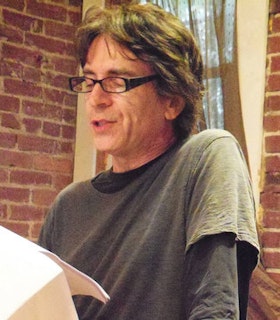 A close-up portrait of Gari Lutz wearing a gray t-shirt, thin black glasses, and reading from a paper in front of a brick wall. She looks down at the paper appearing deep in thought. 