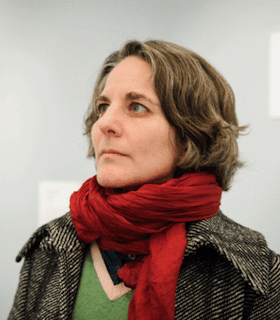 A side angle portrait of Mac Low looking up and towards the distance in front of a white wall. She has short brown hair and wears a green sweater, red scarf, and herringbone coat.