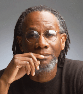 A portrait of Nathaniel Mackey in front of a grey wall. He looks directly at the camera and has his hand lifted by his mouth. He has shoulder length hair and wears wire rimmed glasses and a brown sweater. 