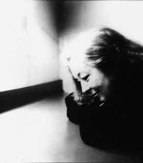 A blurred black and white portrait of Ruth Maleczech leaning on a counter with her elbows, looking down towards it. She wears a black top and has medium length hair. 