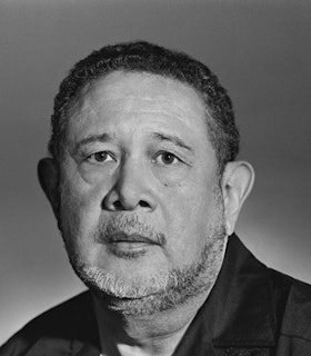 A close up black and white portrait of Jaime Manrique looking directly at the camera. He wears a dark shirt and has closely cut hair. The background is all gray.