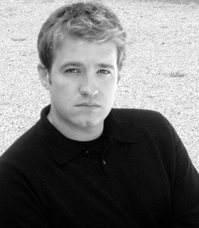 A black and white portrait of Caden Manson looking directly at the camera. He has short hair and wears a black sweater. He appears to be standing in front of a background of gravel.