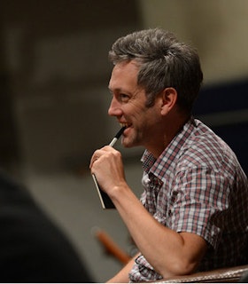 A  profile portrait of Richard Maxwell in front of a blurred background. He smiles and bites the cap of a sharpie as he looks out towards something beyond the frame of the image. He wears a grey and red checkered shirt and has short grey hair. 