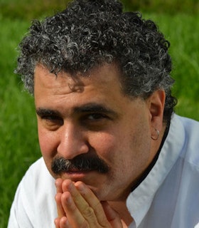 A portrait of Marc Mellits in front of a grassland. He places his hands together under his chin.