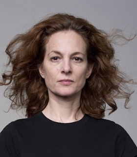 A portrait of Jodi Melnick in front of a light grey background. She has medium length curly auburn hair which is blown out behind her and wears a black shirt. 