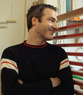 A portrait of Ohad Meromi within a white room, standing next to a red rack. He wears a navy sweater with red detailing. With his arms crossed at his chest, he smiles and looks over his left shoulder. 