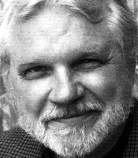 A close-up black and white portrait of Douglas Messerli looking directly at the camera and smiling. He has short hair and a some facial hair and wears a checkered print top.