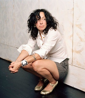 A portrait of Sarah Michelson crouching on the floor in front of a white painted plywood wall. She has black curly hair, a tattoo of a word written in script on her forearm, and wears a white button up shirt, grey striped shorts, a white bracelet with the chanel logo, and gold high heels. 