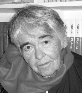 A black and white portrait of Kate Millet in front of a shelf of books written by her. She has short grey hair and wears a grey scarf. 