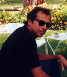 A side angled portrait of Curtis Mitchell in front of a garden with grass, a tree trunk, orange floral bushes and a white table and chair. He has short dark hair and wears black sunglasses and a black short sleeved shirt. 