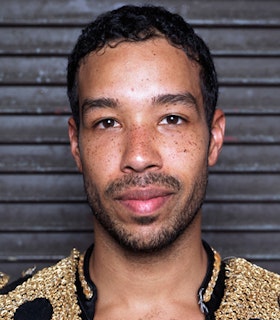 A portrait of Rashaun Mitchell in front of a horizontally striped grey wall. He has short black hair, a short beard, and wears a gold beaded jacket.