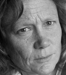 A close-up black and white portrait of Jennifer Monson looking directly at the camera. She has mid-length curly hair and wears a thoughtful expression on her face. 