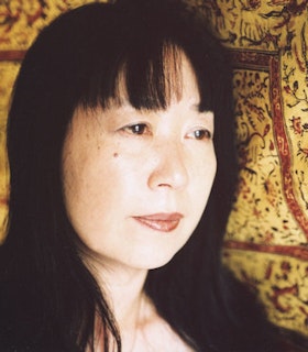 A side portrait of Ikue Mori in front of a patterned backdrop in earthy yellow and terracotta colors. She has dark shoulder-length hair with bangs.