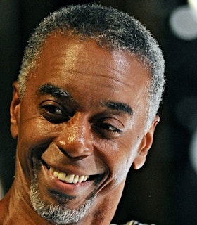 A close up portrait of Dean Moss angling his head slightly and smiling in front of a black background.