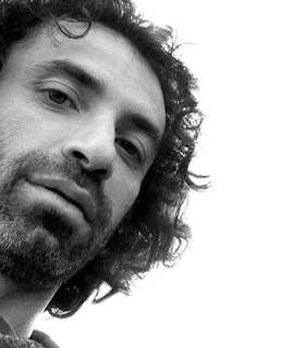 A low angle black and white portrait of Rabih Mroué in front of a bright white background. He has jaw-length curly hair and some facial hair. 
