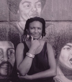 A black and white portrait of Harryette Mullen standing in front of a portrait wall painting. She smiles slightly and supports her chin with her right hand. 