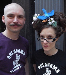 A portrait of Nature Theater of Oklahoma with Pavol Liska on the left and Kelly Copper on the right. Both wear t-shirts which say "Oklahoma" and have a picture of a person riding a horse on them. Pavol Liska is bald and has a handlebar moustache. Kelly Copper wears red lipstick, black glasses, and has her hair in an elaborate updo with white flowers, a monarch butterfly, and a blue bird perched in it. 