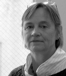 A black and white portrait of Dona Nelson in front of a diamond patterned window. She has short hair and wears thin wire glasses perched atop her head, a white and grey horizontally striped shirt, and a black ruched sweater. 