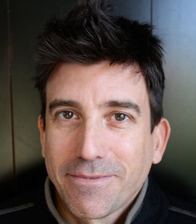 A close up portrait of David Neumann in front of a grey background. He has short black hair, some stubble, and wears a black and light grey high collared shirt.  