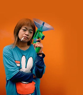 A portrait of Yuko Nexus6 in front of a bright orange background. She has shoulder length orange hair and wears a blue shirt with a cartoon rabbit on it. A cigarette dangles from her lips and she holds a toy green gun in one hand. 