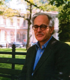 A portrait of Charles North sitting on a bench in front of a grassy plot and a blurred pink building. He has short grey hair and wears brown glasses, a blue shirt, and a grey suit jacket. 