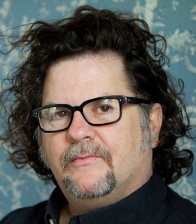 A close up portrait of Tere O'Connor in front of a patterned blue and white background. He has shoulder length curly black hair and a goatee and wears a dark shirt and rectangular black glasses. 