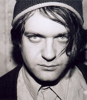 A close up black and white portrait of Jim O'Rourke looking intently at the camera and angling his head downwards. He wears a knit beanie, a striped shirt, and a black cardigan. He has short black hair. The background is blurred and grey.