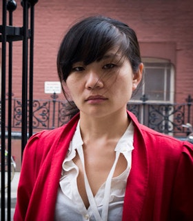 A portrait of Okkyung Lee in front of a red brick building. She has side swept black bangs and the rest of her hair is pulled into a low pony tail. She wears a cherry red jacket and a white shirt. 