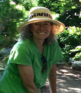 A portrait of Maureen Owen in a place filled with plants. She has shoulder length curly gray hair and wears a straw hat with a zebra print sash around it, and a lime green shirt. She looks directly at the camera and smiles. 