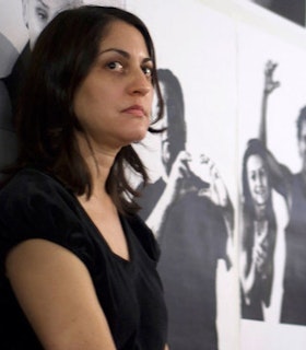 A side angled portrait of Alix Pearlstein in front of a wall printed with life sized photos of people. She has medium length black hair and wears a black short sleeved t-shirt. 