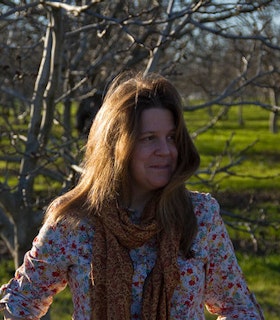A side angled portrait of Katie Peterson in front of a bare tree and grass filled plain. She has medium length light brown hair and wears a multi-colored floral shirt and an orange and red paisley patterned scarf. 
