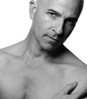 A close up black and white portrait of Stephen Petronio against a white background. Petronio is shirtless and rests one hand against his chest. He looks directly at the camera with a sly expression on his face. 